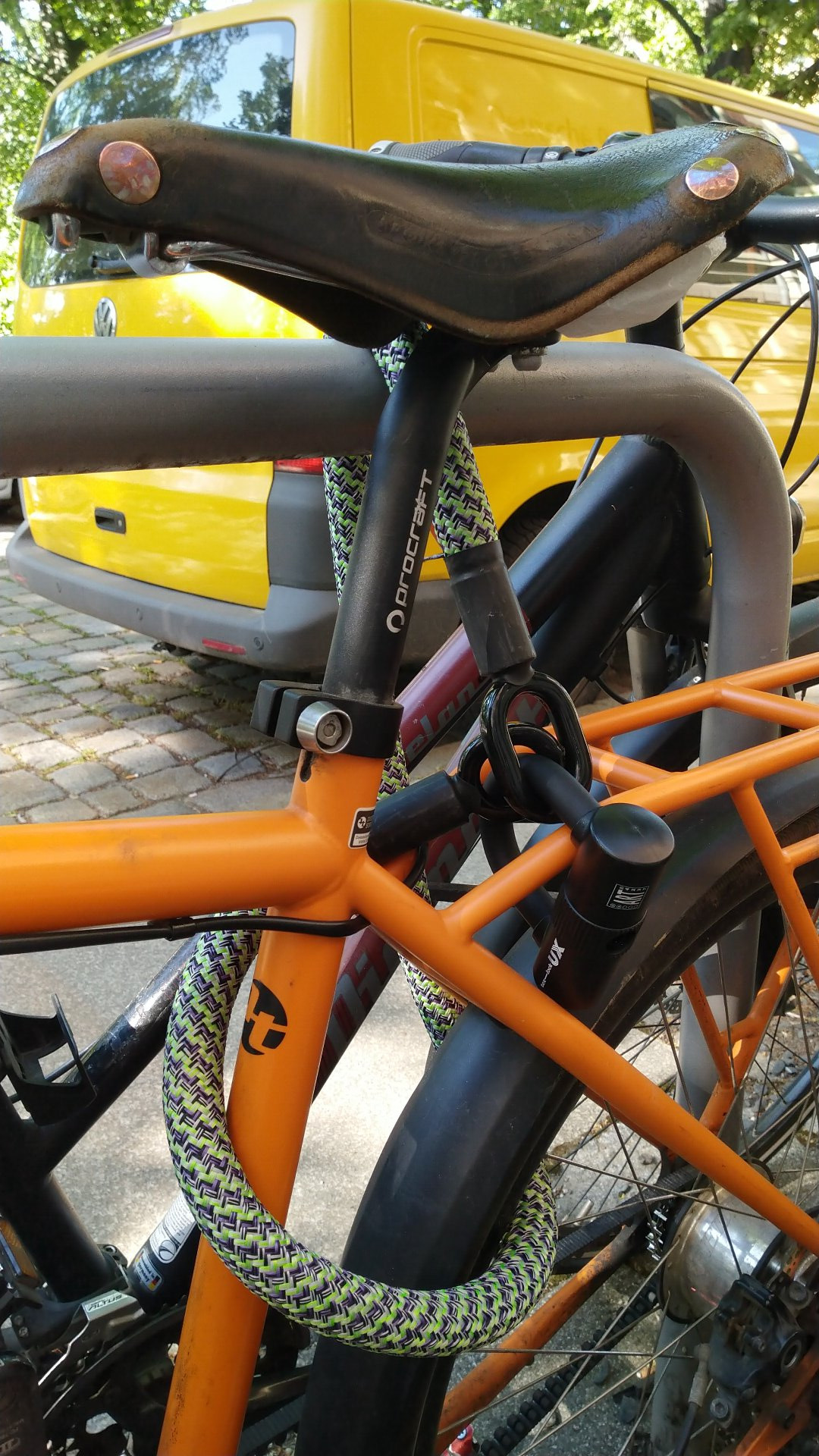 Das tex-lock eyelet in grün/schwarz schließt das organgene Rad an einem Fahrrad-Bügel an. Der kleine Bügel verschließt die beidenen Enden am Gepäckträger des Rads. Der Gepäckträger ist beim Tout Terrain fest mit dem Rahmen verbunden und ebenfalls in orange.